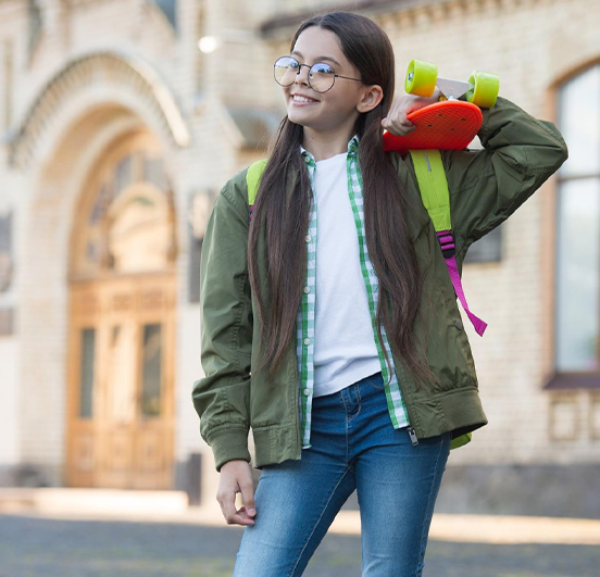 Uma jovem sorri enquanto posa com um skate laranja e amarelo nas costas. Ela usa óculos redondos, uma camiseta branca, uma camisa xadrez, uma jaqueta verde-oliva e jeans. Um prédio de tijolos com arcos é visível ao fundo.