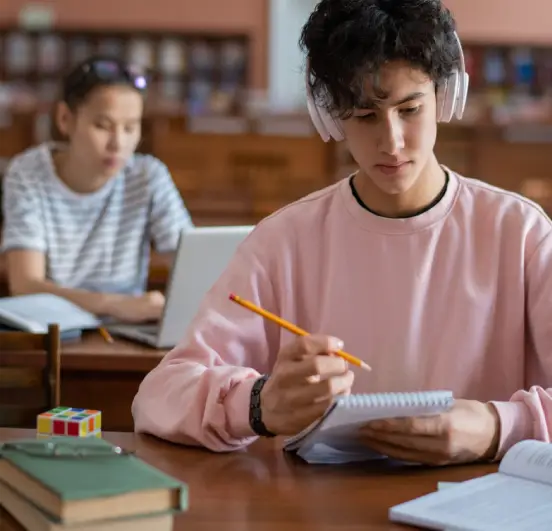 Um estudante do ensino médio usa fones de ouvido brancos enquanto escreve em um caderno. Ele usa um moletom rosa e tem cabelo escuro e encaracolado. Um cubo mágico, livros e uma pilha de papéis estão na mesa à sua frente.