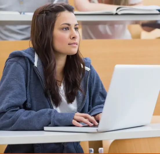 Uma jovem com cabelo castanho comprido está sentada em uma sala de aula, usando uma blusa branca e um moletom azul escuro com capuz. Ela olha para longe enquanto digita em um laptop.