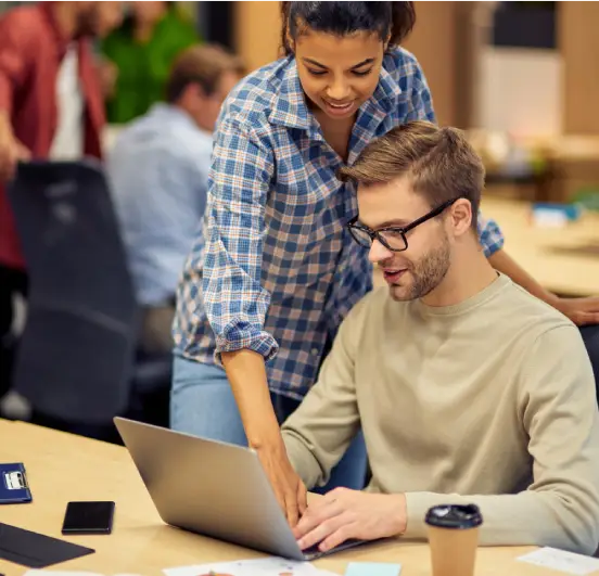 Uma mulher de cabelo escuro e camisa xadrez azul se inclina sobre o ombro de um homem. Ela aponta para seu laptop, onde o homem de óculos e suéter bege está trabalhando.