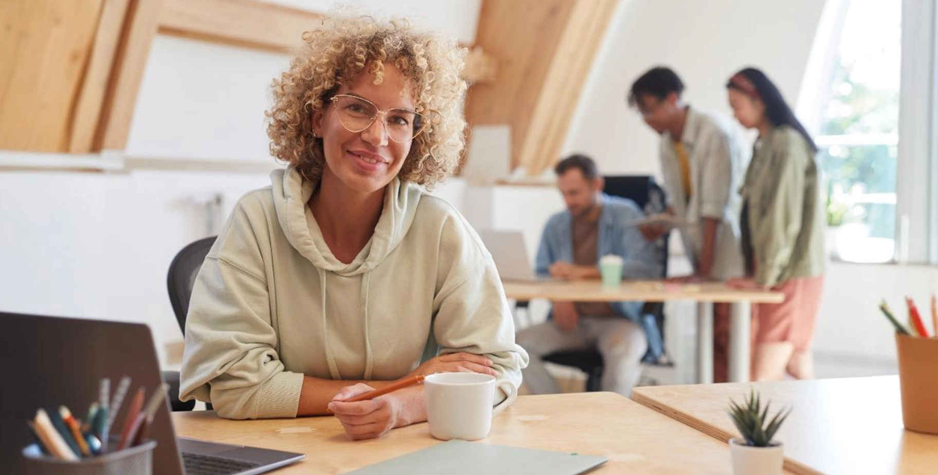 Uma mulher com cabelo cacheado e loiro, usando óculos e um moletom verde claro, senta-se em uma mesa de madeira e sorri para a câmera. Ela segura um lápis na mão direita e tem uma xícara branca e um laptop à sua frente.