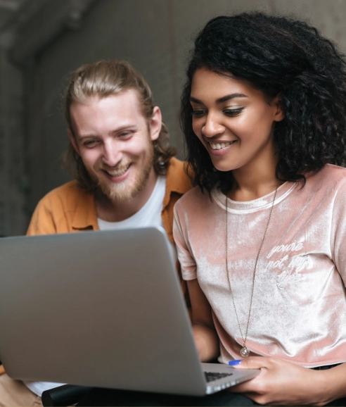 Um homem sorridente de cabelo comprido e loiro, com barba, e uma mulher sorridente de cabelo crespo e escuro olham para a tela de um laptop. O homem usa uma camisa marrom e a mulher, uma blusa rosa de veludo com um colar comprido.