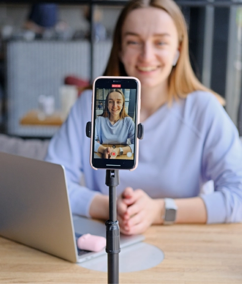 Uma mulher loira sorridente, com fones de ouvido sem fio, senta-se em uma mesa de madeira. Ela usa uma blusa azul clara e um relógio prateado. À frente dela, um smartphone está em um tripé, mostrando a própria mulher em uma chamada de vídeo.