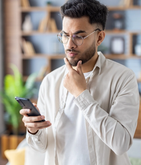 Um homem jovem com barba e óculos redondos segura um smartphone preto na mão esquerda e apoia a mão direita no queixo, com uma expressão pensativa. Ele usa uma camisa de botão clara sobre uma camiseta branca. 