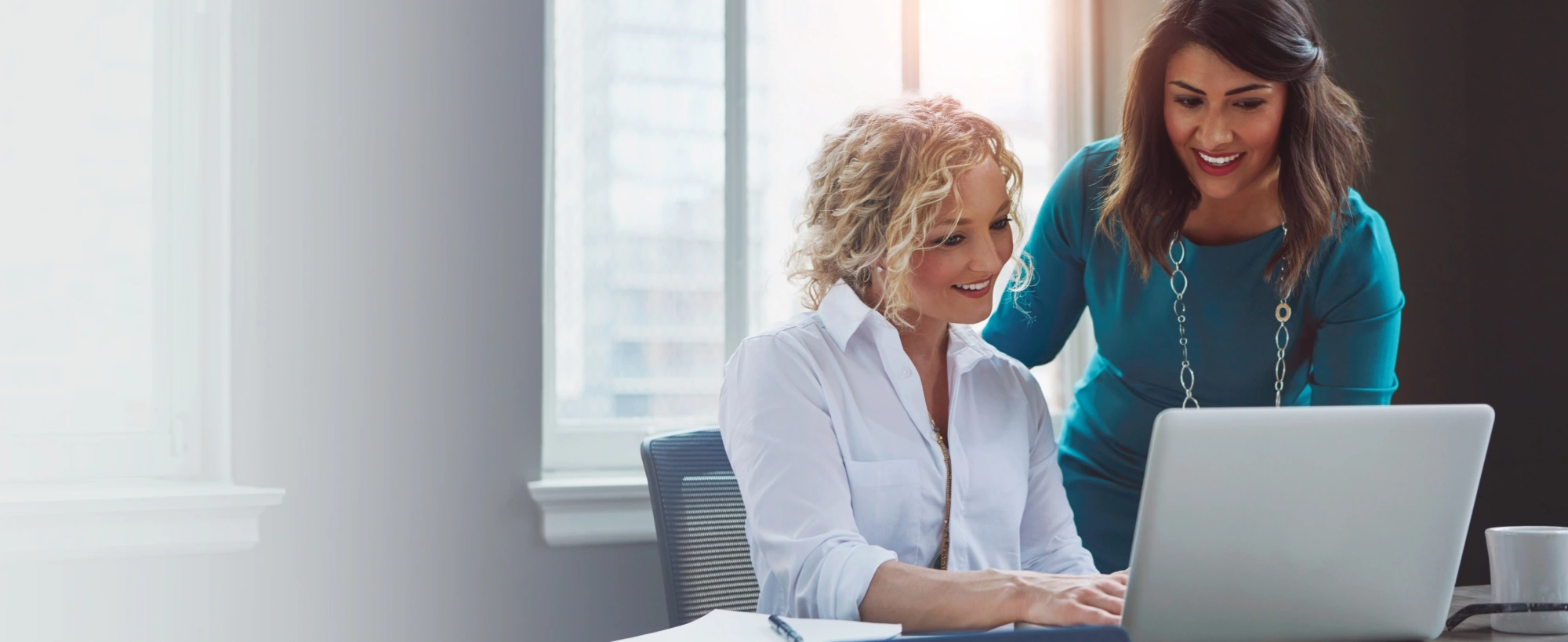 Dois estudantes, um homem e uma mulher, sorriem para a câmera em frente a um prédio universitário. A mulher, à esquerda, usa óculos e segura um caderno. O homem, à direita, usa uma mochila.