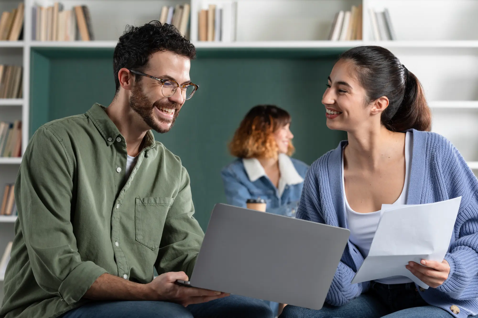 Dois jovens sorrindo enquanto usam um notebook e revisam papéis durante uma sessão de estudos.