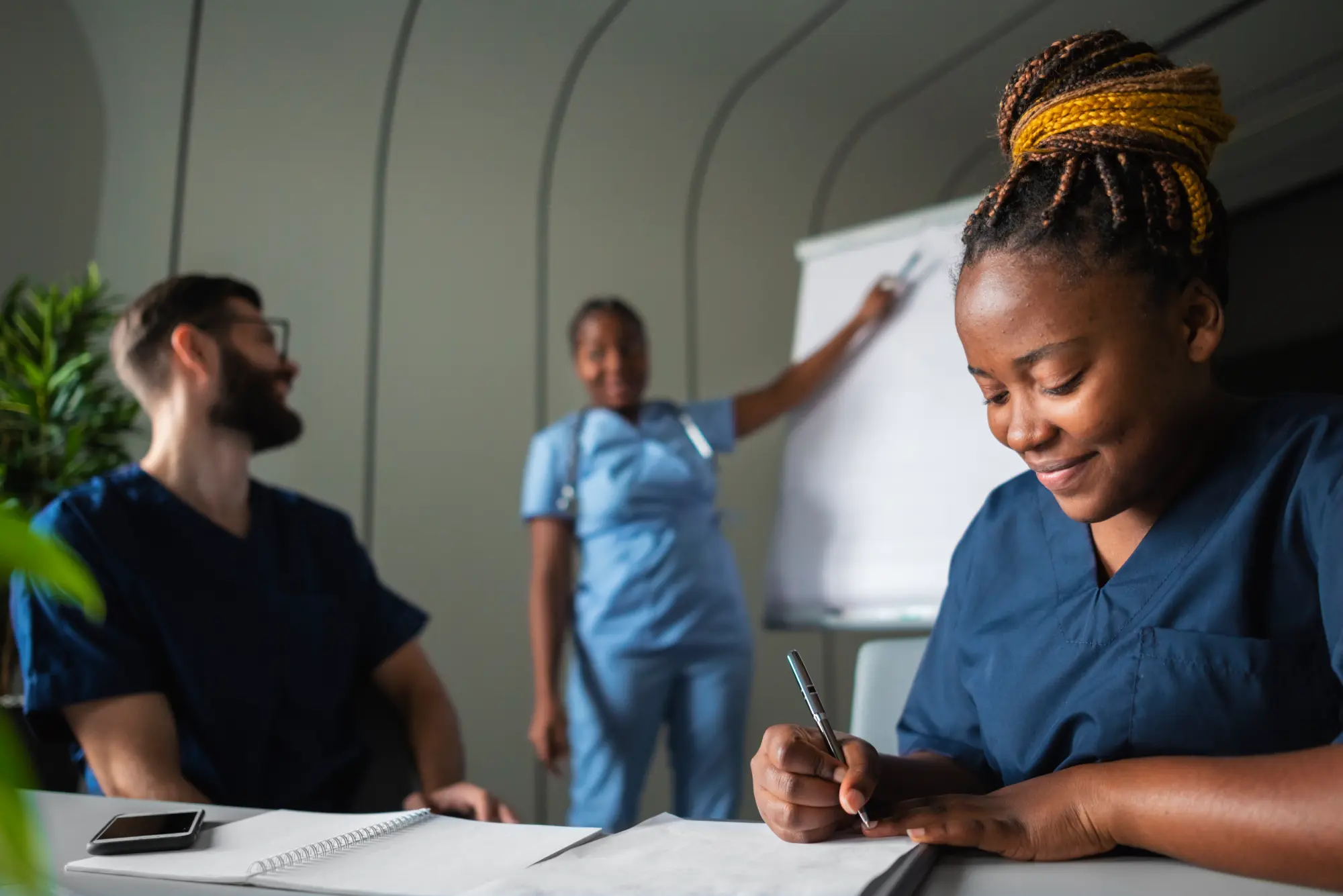 Mulher em uniforme médico fazendo anotações durante uma apresentação em sala de aula.
